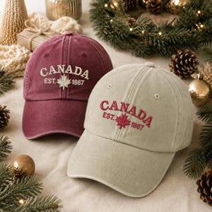 Puede incluir: Dos gorras de béisbol, una granate y otra beige, cada una con el texto bordado CANADA EST. 1867 sobre un icono de hoja de arce. La gorra granate tiene bordados en color crema, mientras que la beige tiene bordados en color rojo oscuro.