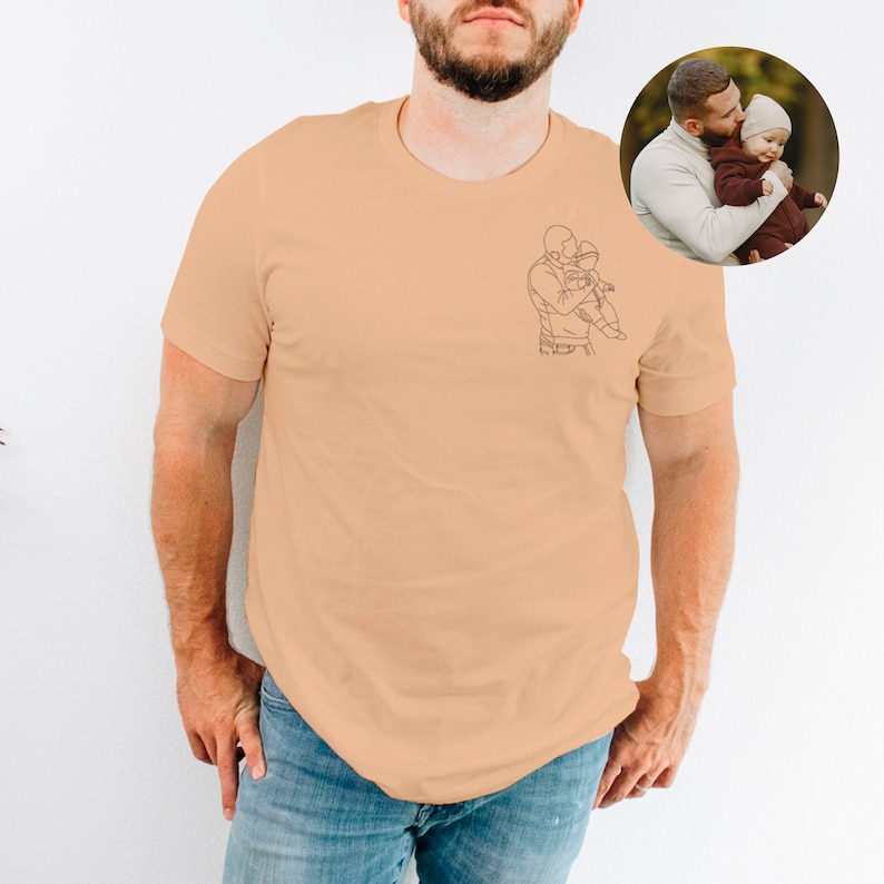 May include: A light peach t-shirt with a line drawing of a father holding a child. The drawing is on the left side of the shirt.