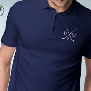 Puede incluir: Polo azul marino con un diseño bordado blanco con temática de golf en el pecho. El diseño incluye palos de golf cruzados, una pelota de golf y las letras "U" y "M". La camisa tiene mangas cortas y cuello.