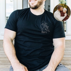 May include: A black t-shirt with a white line drawing of a father and child hugging. The drawing is on the left side of the shirt.