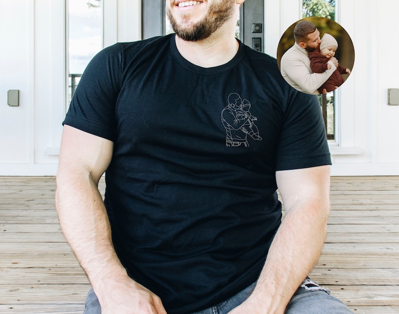 May include: A black t-shirt with a white line drawing of a father and child hugging. The drawing is on the left side of the shirt.