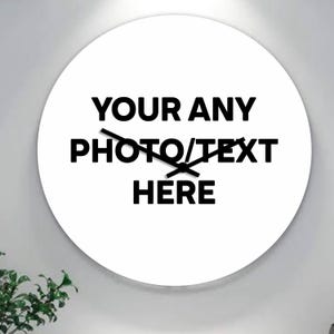 Puede incluir: Un reloj de pared redondo blanco con manecillas negras y el texto "YOUR ANY PHOTO/TEXT HERE" en letras negras en negrita. El reloj está montado en una pared gris claro, con un vistazo de follaje verde en la esquina inferior izquierda.