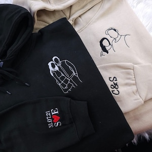 May include: Two black and beige hooded sweatshirts with embroidered designs. One sweatshirt has a couple's silhouette with the text "S&E 12.20.21" and a red heart. The other sweatshirt has a couple's silhouette and the text "C&S".