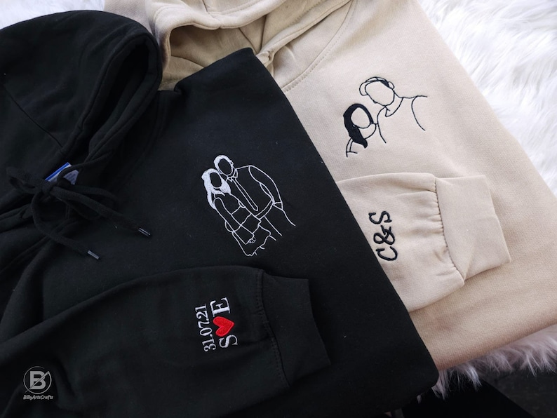 May include: Two black and beige hooded sweatshirts with embroidered designs. One sweatshirt has a couple's silhouette with the text "S&E 12.20.21" and a red heart. The other sweatshirt has a couple's silhouette and the text "C&S".