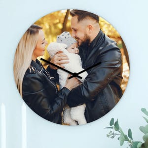 Puede incluir: Reloj de pared redondo con una foto familiar. La esfera del reloj muestra una foto de una familia con un bebé. El reloj tiene manecillas negras y está montado en una pared blanca.
