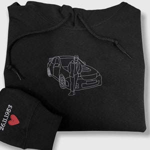 Op de afbeelding: Zwarte hoodie met een witte omtrek van een persoon die naast een auto staat. De hoodie heeft een klein zwart patch met een rood hart en de tekst "26.11.1983" op de linkermouw.