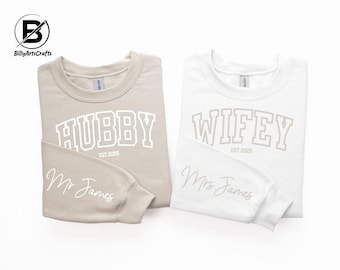 May include: Two beige and white sweatshirts with the words "Hubby Est. 2023" and "Wifey Est. 2023" printed on them. The sweatshirts also have the names "Mr. James" and "Mrs. James" printed on them.