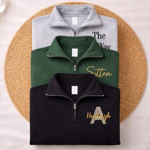 May include: Three quarter-zip sweatshirts in grey, green, and black, stacked on a woven mat. The grey sweatshirt has the text "The Way", the green one says "Sutton", and the black one says "Huxleigh" with a letter "A".