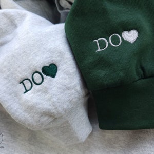 May include: Two hooded sweatshirts with the word "DO" embroidered on them. One sweatshirt is grey with a green heart and the other is green with a white heart.