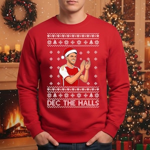 May include: Red Christmas sweater with a festive pattern of snowflakes and trees. The sweater features a graphic of a person wearing a Santa hat and clapping. The text "DEC THE HALLS" is displayed on the sweater.