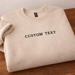 May include: A beige sweatshirt with the words "CUSTOM TEXT" embroidered in black. The sweatshirt is folded on a wooden surface. The garment has a crew neck and long sleeves.