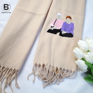 May include: Beige scarf with an embroidered image of two people sitting together. The scarf has fringe on the bottom and the embroidery is in shades of pink, purple, and black.