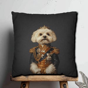 Puede incluir: Cojín decorativo con un perro maltés real con uniforme militar y corona dorada. El perro está sobre un fondo gris oscuro. El cojín tiene una funda negra y está colocado sobre una superficie de madera.