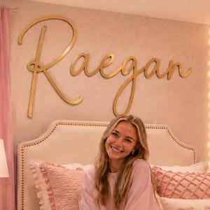 May include: A bedroom featuring a cream-coloured upholstered headboard with gold nailhead trim, a pink textured comforter, and a wall-mounted gold script sign reading Raegan. The room includes a white cube shelf, a pink ottoman, and a Matisse print.