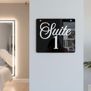 Pode incluir: Uma placa quadrada preta com as palavras "Suite 1" em branco. A placa está montada numa parede branca. A imagem é tirada em ambientes fechados, com um espelho e uma planta visíveis no fundo.