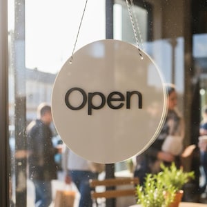 Puede incluir: Un letrero redondo blanco con la palabra "Open" en letras negras cuelga en un escaparate. La imagen muestra una escena de café al aire libre soleada con mesas, plantas y personas que pasan.