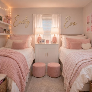 Op de afbeelding: Twee eenpersoonsbedden met wit beddengoed en roze textuurrijke plaids, voorzien van houten wandborden met de namen Emily en Eva. De kamer bevat twee roze fluwelen poefs, een witte kast en twee tafellampen met roze voet en witte kap.