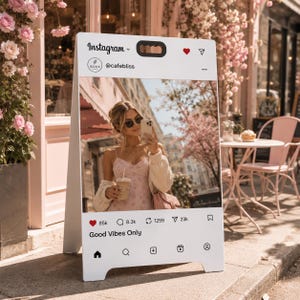 Könnte beinhalten: Ein weißer A-förmiger Gehwegaufsteller mit einer aufgedruckten Instagram-Oberfläche und dem Benutzernamen @cafebliss. Das Display zeigt ein Foto einer Person in einem Blumenkleid und Strickjacke mit Getränk sowie den Text Good Vibes Only.