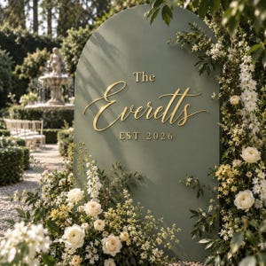 May include: A sage green arched sign featuring gold lettering that reads The Everetts EST 2026. The sign is decorated with white roses and greenery, positioned upright in an outdoor setting with a fountain and chairs visible in the background.