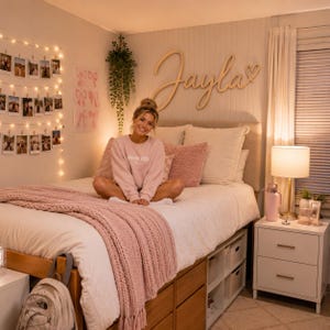 Op de afbeelding: Een houten wandbord met de naam Jayla in cursief schrift en een hartje, gemonteerd boven een bed met wit beddengoed, een roze gebreide deken, roze kussens en een lichtsnoer met foto's aan de muur.