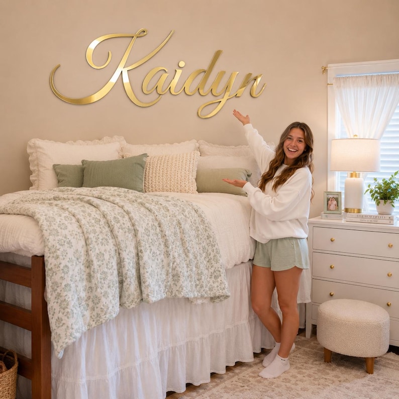 Op de afbeelding: Een groot, goudkleurig wandbord in scriptstijl met de naam Kaidyn, gemonteerd op een beige muur boven een bed. Het bord heeft een reflecterende, metalen afwerking en elegante, vloeiende cursieve letters die horizontaal over de muur lopen.