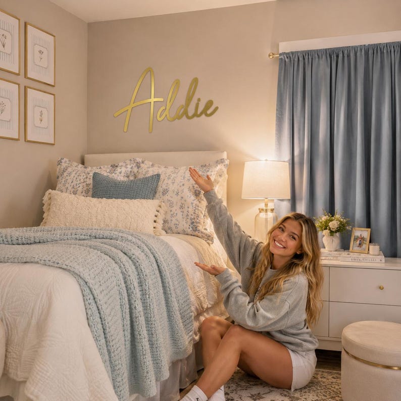 Op de afbeelding: Een goudkleurig metalen wandbord met de naam Addie in een cursief lettertype, gemonteerd op een beige muur boven een bed met blauw en wit beddengoed, een textuurrijke sprei en bijpassende blauwe gordijnen.