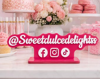 May include: A bright pink acrylic social media sign featuring the white text @Sweetdulcedelightss and white circular icons for Facebook, Instagram, and TikTok. The sign has a wavy top edge and stands on a rectangular base, designed for display.