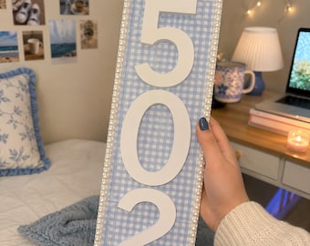 Op de afbeelding: Een rechthoekig verticaal bord met een lichtblauwe en witte gingham-achtergrond en de grote witte cijfers 502 in het midden. De randen van het bord zijn versierd met een boord van kleine witte kunstparels.