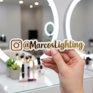Puede incluir: Una pegatina blanca con letras doradas y el logotipo de Instagram. El texto dice "@MarcosLighting". La pegatina se sostiene frente a un tocador con brochas de maquillaje y un espejo.