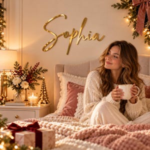 Op de afbeelding: Een goudkleurig wandbord met de naam Sophia in een cursief lettertype. De letters zijn bevestigd op een effen muur boven een bed en hebben een metallic afwerking die het licht in de slaapkamer reflecteert.