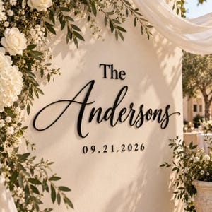 May include: A wall-mounted sign featuring black, three-dimensional lettering that reads The Andersons and the date 09.21.2026. The sign is displayed on a smooth, light-coloured surface, accented by white roses and green foliage on the left side.