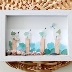 Puede incluir: Una obra de arte enmarcada con temática de playa que presenta cuatro pájaros estilizados hechos de vidrio marino posados en postes de madera. La obra incluye vidrio marino azul, conchas marinas y arena, todo sobre un fondo blanco dentro de un marco blanco.
