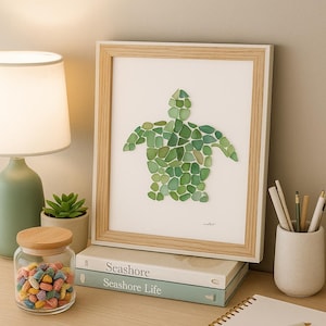 Può includere: Un'opera d'arte incorniciata con un design di tartaruga marina realizzato con vetro marino verde. L'opera è esposta su una scrivania con libri, una lampada, un barattolo di caramelle colorate e un blocco note. La cornice è in legno chiaro.