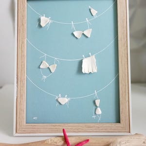 Könnte beinhalten: Gerahmtes Kunstwerk mit hellblauem Hintergrund, das weiße Linien als Wäscheleinen zeigt. Weiße, geformte Bikini-Oberteile und -Unterteile sind an den Leinen befestigt. Der Rahmen ist hellbraun. Ein roter Seestern und Treibholz befinden sich im Vordergrund.