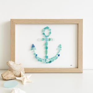 Puede incluir: Una obra de arte enmarcada con un diseño de ancla hecho de vidrio marino en tonos azules y verdes. El ancla está sobre un fondo blanco dentro de un marco de madera natural. Conchas marinas decorativas y madera flotante cerca del marco.
