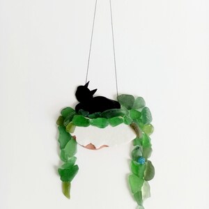 Peut inclure: Une silhouette de chat noir suspendue à une ficelle, entourée de morceaux de verre vert et bleu, formant un pot de plante.