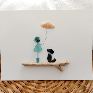 Peut inclure: Une œuvre d'art fantaisiste représentant une silhouette et un chien faits de verre de mer et de galets. La silhouette, vêtue d'une robe et d'un chapeau bleus, tient un parapluie en coquillage. Le chien porte un collier bleu, le tout sur un perchoir en bois flotté.