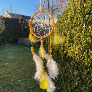 Peut inclure: Un attrape-rêves jaune avec des plumes blanches et des perles en bois qui pendent. L'attrape-rêves est suspendu à un support métallique dans un jardin.