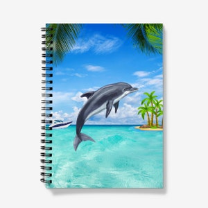 Könnte beinhalten: Ein spiralgebundenes Notizbuch mit einem bunten Cover, das einen Delfin zeigt, der aus dem Wasser springt, Palmen und einen blauen Himmel.