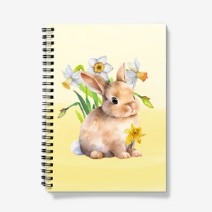Könnte beinhalten: Ein spiralgebundenes Notizbuch mit einer Aquarellillustration eines braunen Hasen und weißen und gelben Narzissen. Der Hase hält eine gelbe Blume. Der Hintergrund ist ein helles Gelb.