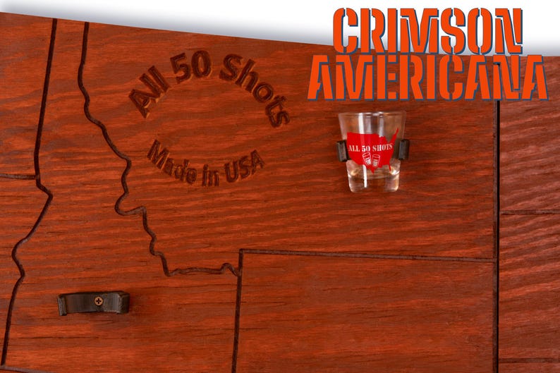 K&ouml;nnte beinhalten: Eine Holzkarte der Vereinigten Staaten mit dem Text "All 50 Shots Made in USA" und "Crimson Americana" darauf gedruckt. Ein Schnapsglas mit dem Text "All 50 Shots" ist an der Karte befestigt.