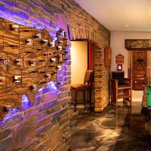 K&ouml;nnte beinhalten: Eine Holzkarte der Vereinigten Staaten mit einer blauen Lichtumrandung, die an einer Backsteinwand montiert ist. Die Karte ist mit kleinen Schnapsgl&auml;sern verziert, die jeweils einen anderen Bundesstaat repr&auml;sentieren. Der Raum verf&uuml;gt &uuml;ber einen Billardtisch mit gr&uuml;nem Filz und einer Buntglasleuchte.