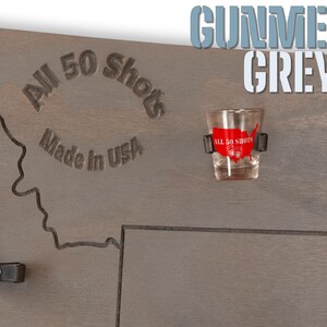 K&ouml;nnte beinhalten: Ein Holz-Shotglas-Halter mit einer Karte der Vereinigten Staaten, auf der die Worte "All 50 Shots" und "Made in USA" eingraviert sind. Der Halter ist in Gunmetal Grey lackiert und enth&auml;lt ein Shotglas mit einem roten und wei&szlig;en "All 50 Shots" Aufkleber.