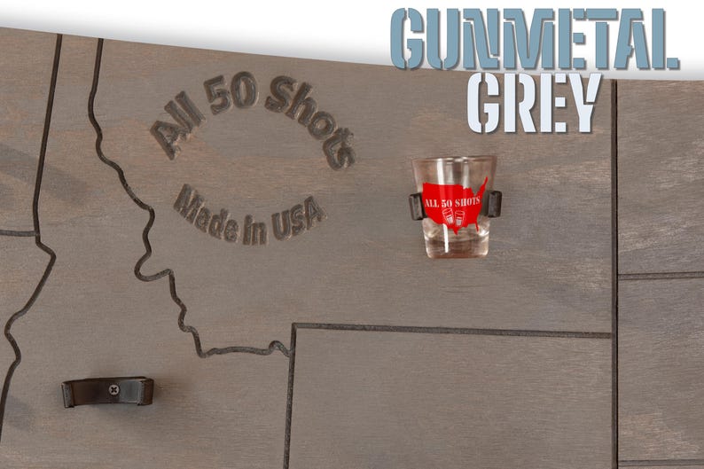 K&ouml;nnte beinhalten: Ein Holz-Shotglas-Halter mit einer Karte der Vereinigten Staaten, auf der die Worte "All 50 Shots" und "Made in USA" eingraviert sind. Der Halter ist in Gunmetal Grey lackiert und enth&auml;lt ein Shotglas mit einem roten und wei&szlig;en "All 50 Shots" Aufkleber.