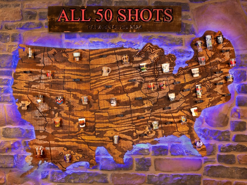 K&ouml;nnte beinhalten: Eine Holzkarte der Vereinigten Staaten mit einem Schnapsglas, das jeden Bundesstaat repr&auml;sentiert. Die Karte ist an einer Backsteinwand befestigt und tr&auml;gt ein Schild dar&uuml;ber mit der Aufschrift "ALL 50 SHOTS ESTABLISHED 2022".