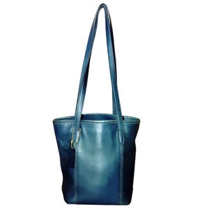 May include: A dark blue leather tote bag with long shoulder straps. The bag has a rectangular shape with a rounded top and a small tag hanging from the handle. The leather appears to be slightly worn, giving it a vintage look.