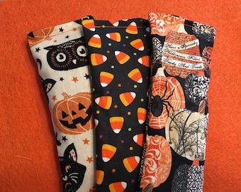 May include: Three Halloween-themed fabric eye pillows. The first pillow has a white background with black owls, pumpkins, and cats. The second pillow has a black background with orange and white candy corn. The third pillow has a black background with orange pumpkins, spiders, and a vintage-style design.