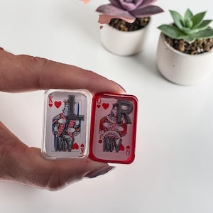 May include: Two rectangular playing card-themed trinkets. One is clear with a King of Hearts design and the letter "L". The other is red with a Queen of Hearts design and the letter "R". Both have the initials "MV".