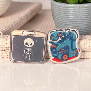 May include: Two square-shaped pins. One features a cartoon skeleton on a gray background with the text "XRAYQUALITY.COM". The other pin has a blue and orange robot design with the text "ODX REVOLUTION".