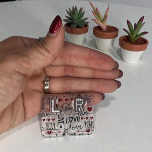 Puede incluir: Iniciales LR de acrílico transparente con temática de amor. El diseño incluye corazones, flechas y la palabra "love" en varias fuentes. Las letras son plateadas. El fondo presenta un patrón repetido de corazones y la palabra "love".